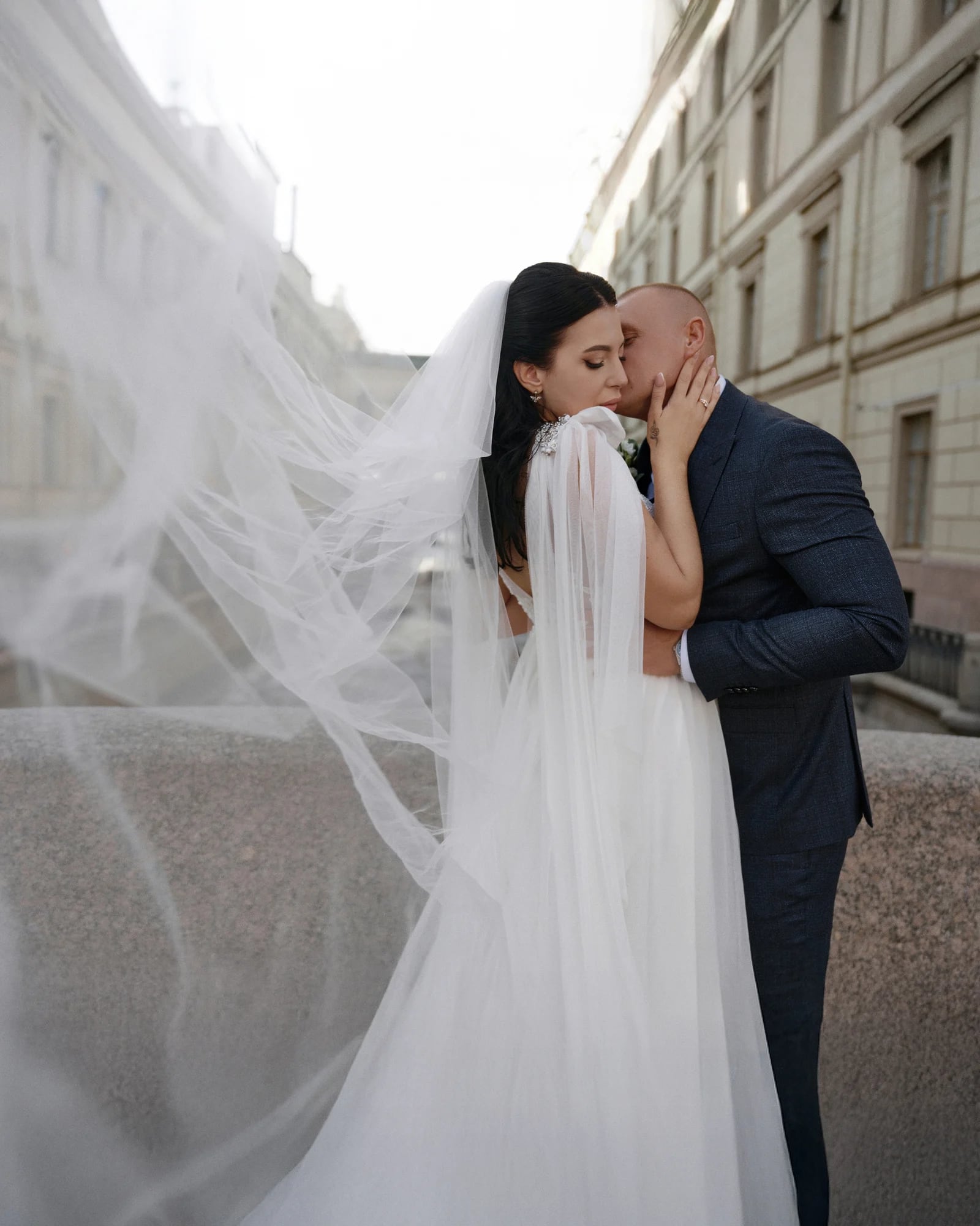 Невеста и жених обнимаются на улице, фата развевается на ветру