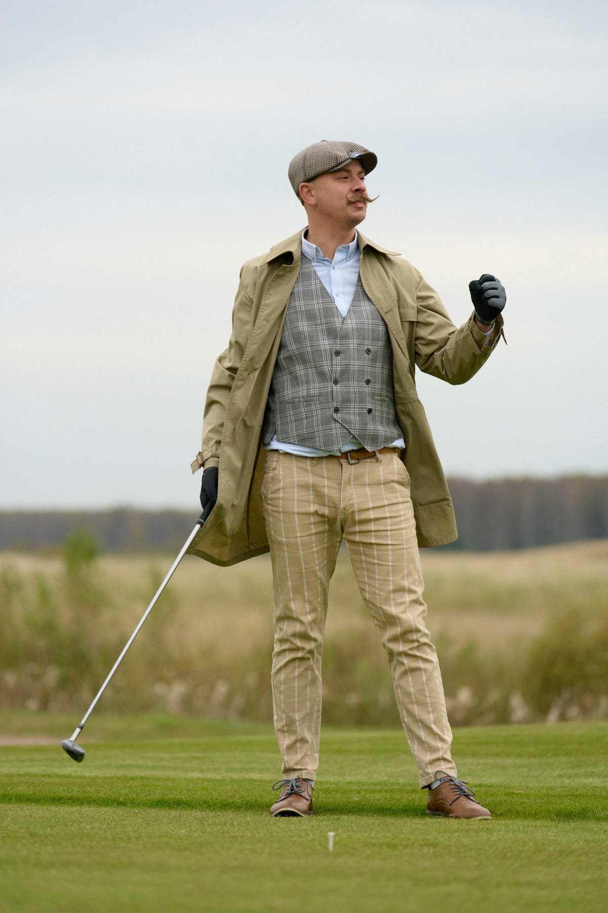 Гольфист в клетчатом костюме, жилете и кепке стоит с клюшкой на поле.