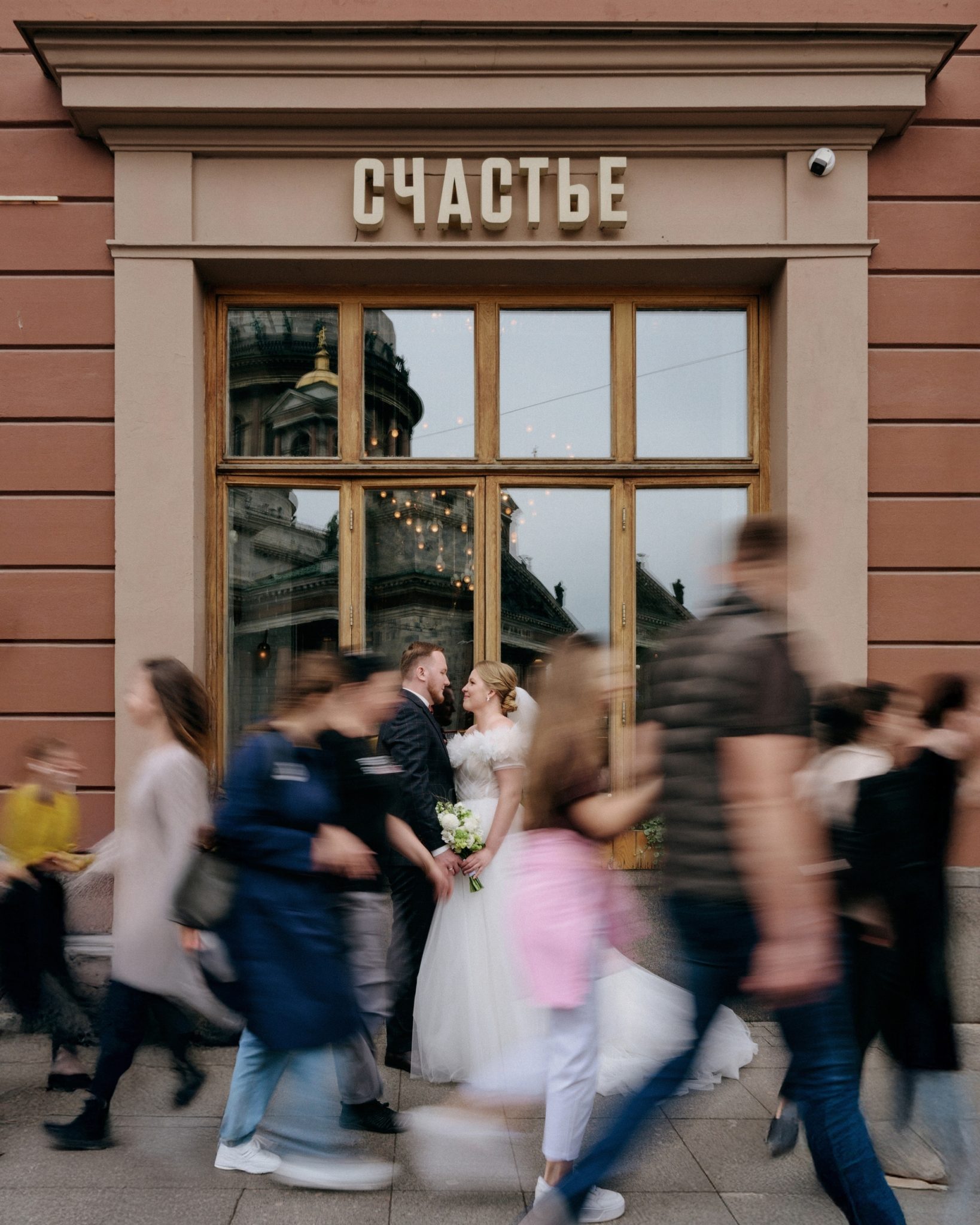 Молодожёны стоят у окна кафе, мимо идут прохожие